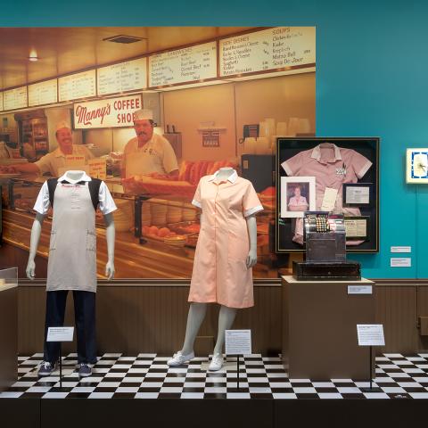 an exhibit of a deli setting with a checkerboard floor and 2 mannequins with deli uniforms on. A large photo behind of a deli counter.
