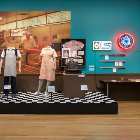 Two mannequins with deli uniforms on with a large photo of deli counter behind them on a platform with a checker board floor.