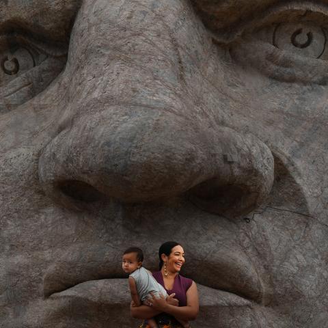 A woman holds a young child in front of a large stone sculpture of a human face.