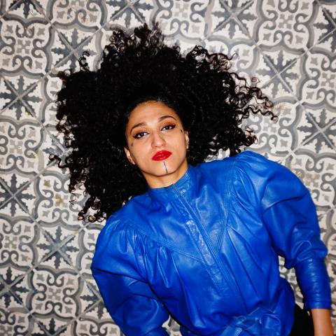 Yousra Mansour, lead singer of the band Bab L'Bluz, lies on a decorative tiled floor wearing a bright blue top. Her hair fans out around her head as she looks directly to the camera.