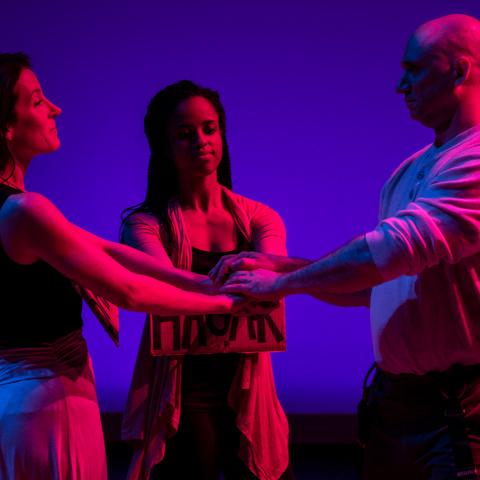 Three people stand on a stage facing each other and holding hand. They are lit with blue and red lights for a performance.