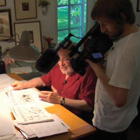 A man with a large camera stands over Maurice Sendak at his desk displaying his illustrations.