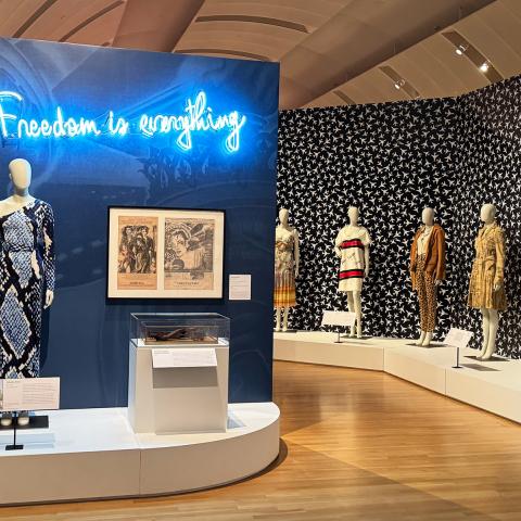 A wide gallery photo showing mannequins in dresses and a neon sign in blue that says Freedom is Everything.