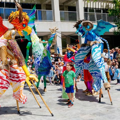 Performers on stilts in elaborate costumes move and dance in the middle of a large group of families in a Courtyard.