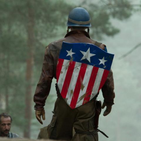 Still from the film showing Captain America walking towards a group of soldiers in a field wearing the iconic Stars and Stripes shield on his back.