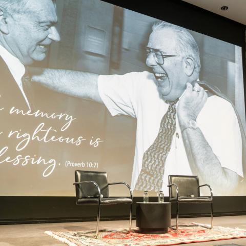 A colorful comic book cover showing Superman flying above a city. A stage is set up with two chairs on a rug with microphones for a discussion with a screen showing Uri Herscher and Howard Friedman.