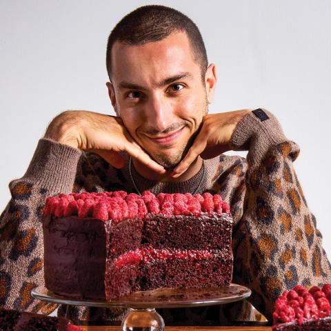 Headshot of Jake Cohen smiling at the camera with his head resting on his hands behind a table with a large, chocolate cake with raspberries. On the right is the cover of the book on a raspberry red color.