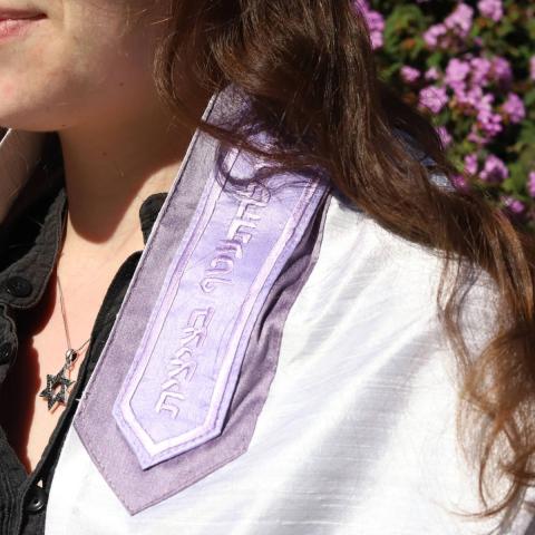 A woman wearing a purple tallit and silver Star of David necklace standing outside in front of a bush of purple flowers.