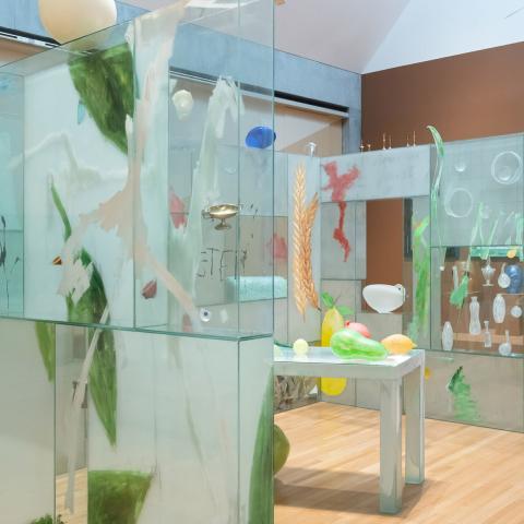 Three glass walls surround a table with colorful objects. Above the walls, larger-then-life feathers suspend from the ceiling up into a skylight.