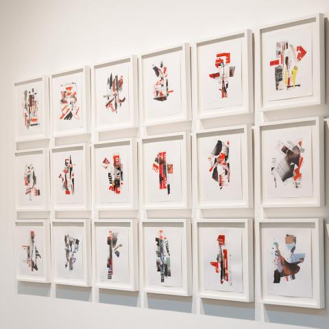 Photo of a gallery corner with a flat screen displaying a colorful video next to 18 framed collages hung in a grid.