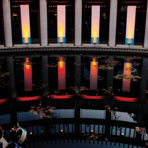 Photo of the lily pond at the Skirball lit up with large column spotlights mimicking a Hanukkah. Families stand along the pond looking at the lights.