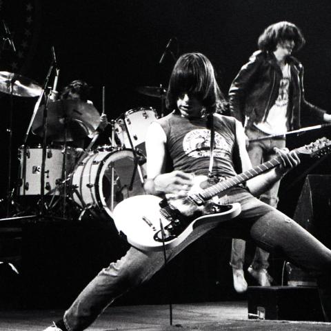 A black and white photo of a guitar player up front from his band on a stage playing.