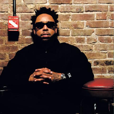 A man with sunglasses sits in front of a brick wall, and a woman stands in front of a harm.