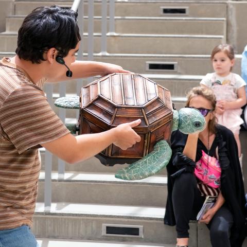A man performs with a turtle puppet in front of families in an outdoor theater.