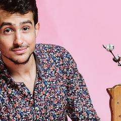 Headshot of Guy Mintus sitting in front of a pink background and surrounded by various, whimsical toys and items