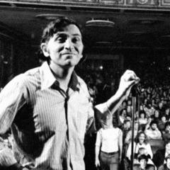 a man on a stage holding a microphone stand with a large crowd of people in the seats below the stage