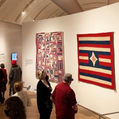 Visitors walking through the museum gallery looking at three quilts with American flag motifs in red, white, and blue.