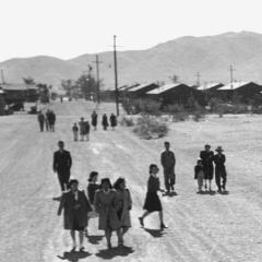 people walking on a dirt road with barracks visible behind them in the distance and mountains even further in the distance