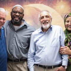Photo of four people posing in front of a colorful backdrop. Three men and a women wearing a headscarf are all holding each other and smiling at the camera