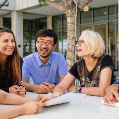 A photo of five people sitting together at a table outside. They are holding papers and pens while laughing with each other.