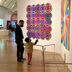 Photo of a museum gallery. Large, colorful quilts are hung on white walls and a myriad of people are looking at them. In the center of the photograph are a father and young son looking at a quilt together.