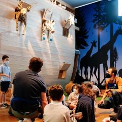 Kids gathered, sitting on the floor with attention on one child speaking. In the noah's Ark rainbow gallery with a wooden ark ing the background and animals hanging of the ark.