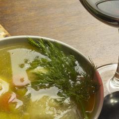 Overhead photo of a bowl filled with matzo ball soup. Next to the bowl is an unleavened cracker, glass of red wine, and spoon.
