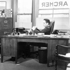 Black and white image of two men sitting at desks in an office conversing. The name "Archer" is painted on the windows behind them.