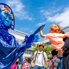 Outdoor photo at the Skirball campus. A large, blue puppet is on the left, reaching out to a young child in their adult's arms and is reaching back. People can be seen behind the trio dancing.