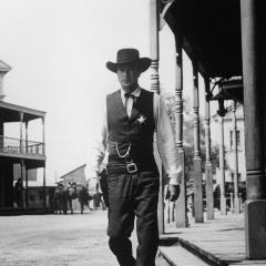 Still from the movie showing Gary Cooper walking towards the camera wearing dark clothing and a cowboy hat. He is surrounded by western buildings and a bright sky.
