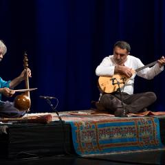 Photo of a dark stage. Kayhan Kalhor and Erdal Erzincan sit on a rug with instruments playing together.