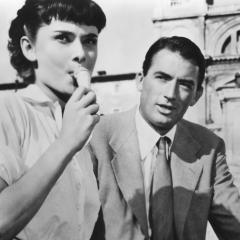 Still from the film Roman Holiday showing Audrey Hepburn and Gregory Peck outside on a street in Rome. Hepburn is eating ice cream, and Peck is looking at her.