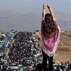 Woman standing on a car with arms up high, looking a the line of stopped cars that goes beyond the horizon. The landscape of Iran beyond.