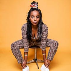 The artist sitting on a stool in front of a yellow backdrop. They are looking directly to camera.