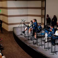 Photo of a group of students lined up on a stage. One student is standing in front of a microphone speaking to the audience.