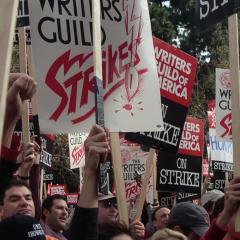 A crowd of people standing together holding signs that read Writers Guild of America On Strike