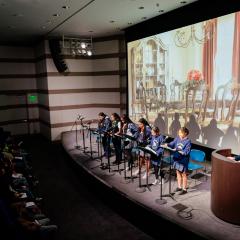 Photo of a group of students lined up on a stage with microphones and stands presenting to a room of people.