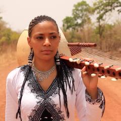 Colorful photograph of Sona Jobarteh standing on a dirt road lined with trees. She is holding her kora over her shoulder and looking slightly away from the camera.