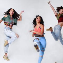 Photo of five ladies smiling and dancing in front of a white background. They are wearing colorful tops, jeans, and gold shoes.