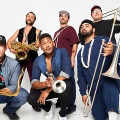 Photo of the six band members standing and kneeling in front of a white background. Each member is holding their instrument.