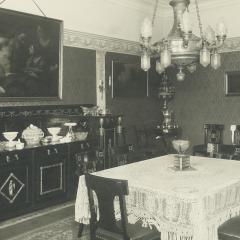Black and white photo of a dining room with a large table and chairs in the center. On the far wall, a large painting is seen hanging.