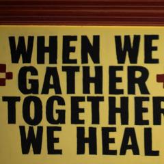 Photo of an outdoor artwork with the words, When We Gather Together We Heal, stamped in black letters on a yellow box.