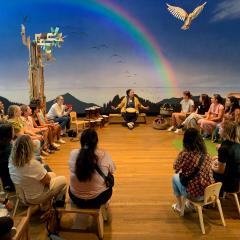 A group of people sitting on chairs in a circle. There is a wood floor. The blue wall in the back of the room has a rainbow painted on it.