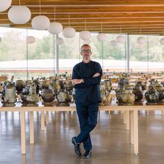 A man standing with arms crossed in front of large table in a large windowed room. The table has row and rows of pottery vessel on it.