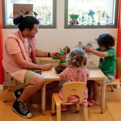 An educator sit with a young boy and girl engaged in making a flower with pipe cleaners and beads in a brightly colored art studio with small tables and orange walls.