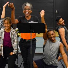 A group of six performers stand in a line posing in song facing foward.