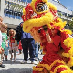 In a large, outdoor crowd, two young girls stand before a performer operating a large, red and yellow, dragon puppet