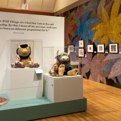 A wall covered in large, colorful flower illustrations and framed artworks appears on the right. On the left is a display of stuffed characters from the book Where the Wild Things Are and a wall quote by the artist Maurice Sendak.
