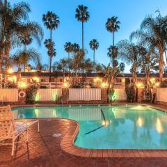 Outdoor swimming pool area with lounge chairs, surrounded by palm trees and lit up by warm lights. A two-story building is visible in the background.