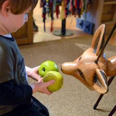 A young child gives two green toy apples to a puppet of a fox.